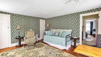 A room with floral wallpaper, a bed with a blue and white bedspread, a chair, a table, and a floor lamp.