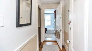 Un couloir aux murs blancs, avec un miroir et une porte donnant sur une salle de bain équipée d’une baignoire et d’une fenêtre.