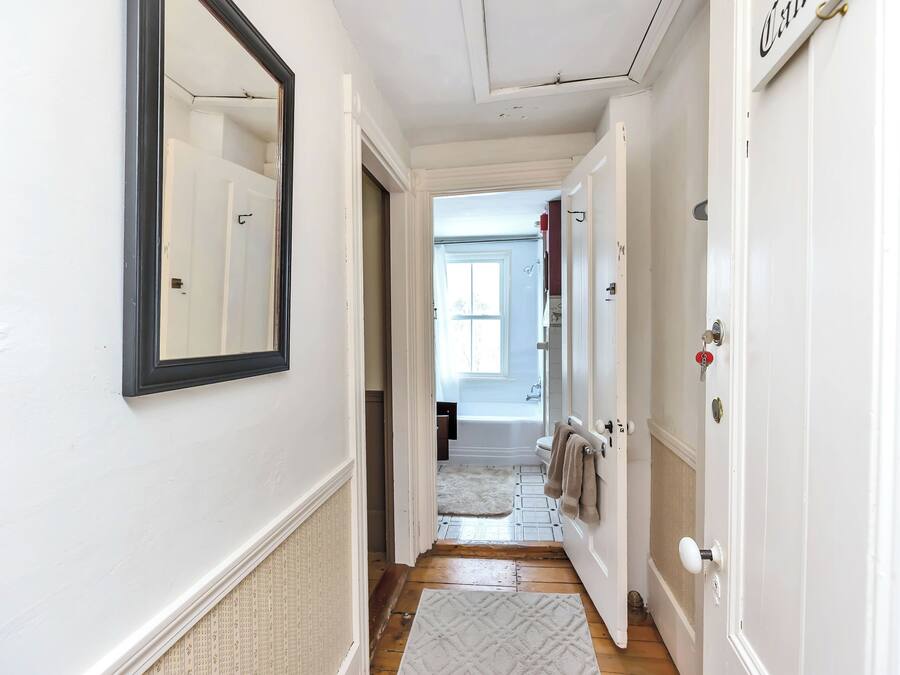 A hallway with white walls, a mirror, and a door leading to a bathroom with a bathtub and a window.