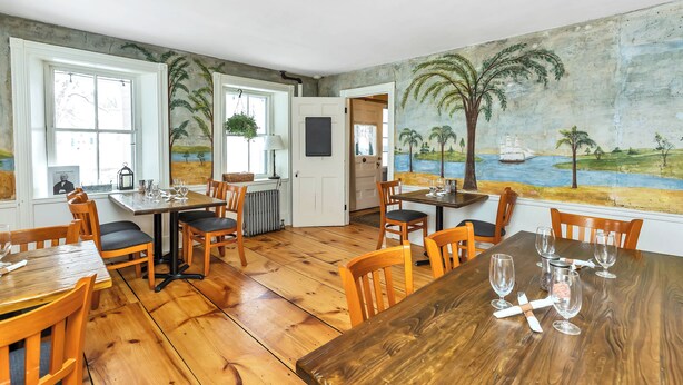 Un espace repas avec des tables et des chaises en bois, une fresque murale et de grandes fenêtres.