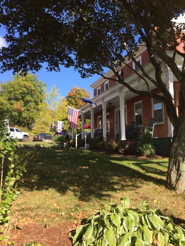 A brick house with a porch, an American flag, and a lawn with fallen leaves.