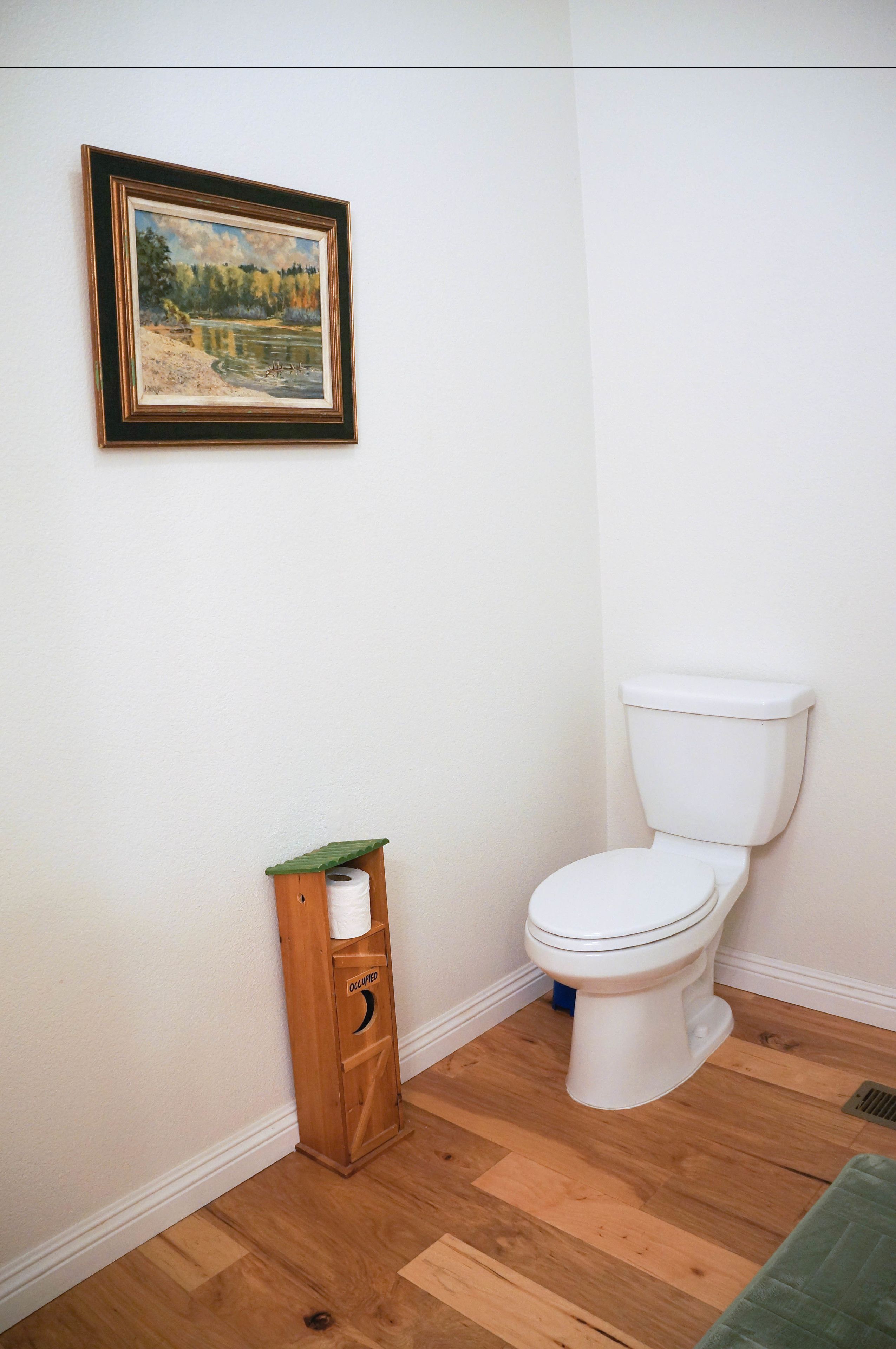 Un baño con inodoro blanco, un dispensador de papel higiénico de madera y un cuadro en la pared.