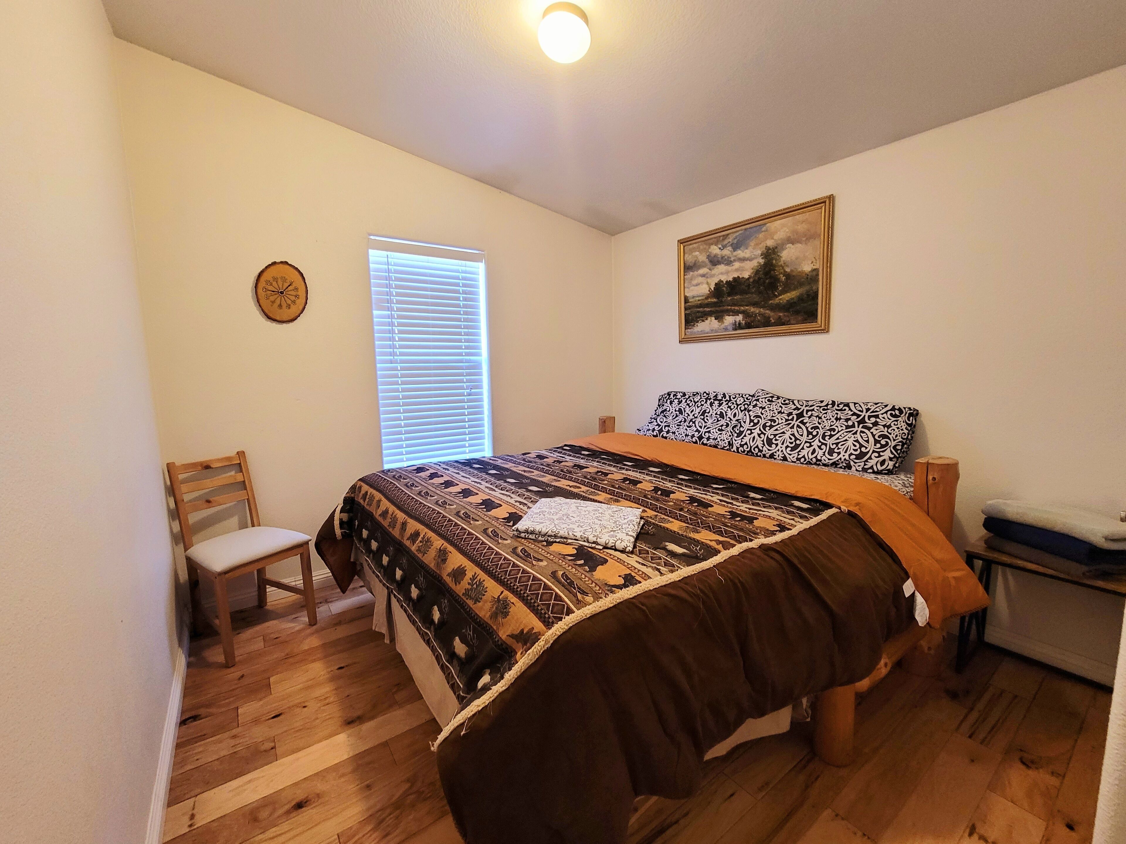 Un dormitorio con suelo de madera, una cama con colcha estampada, una silla, un ventanal con persianas, un reloj y un cuadro en la pared.