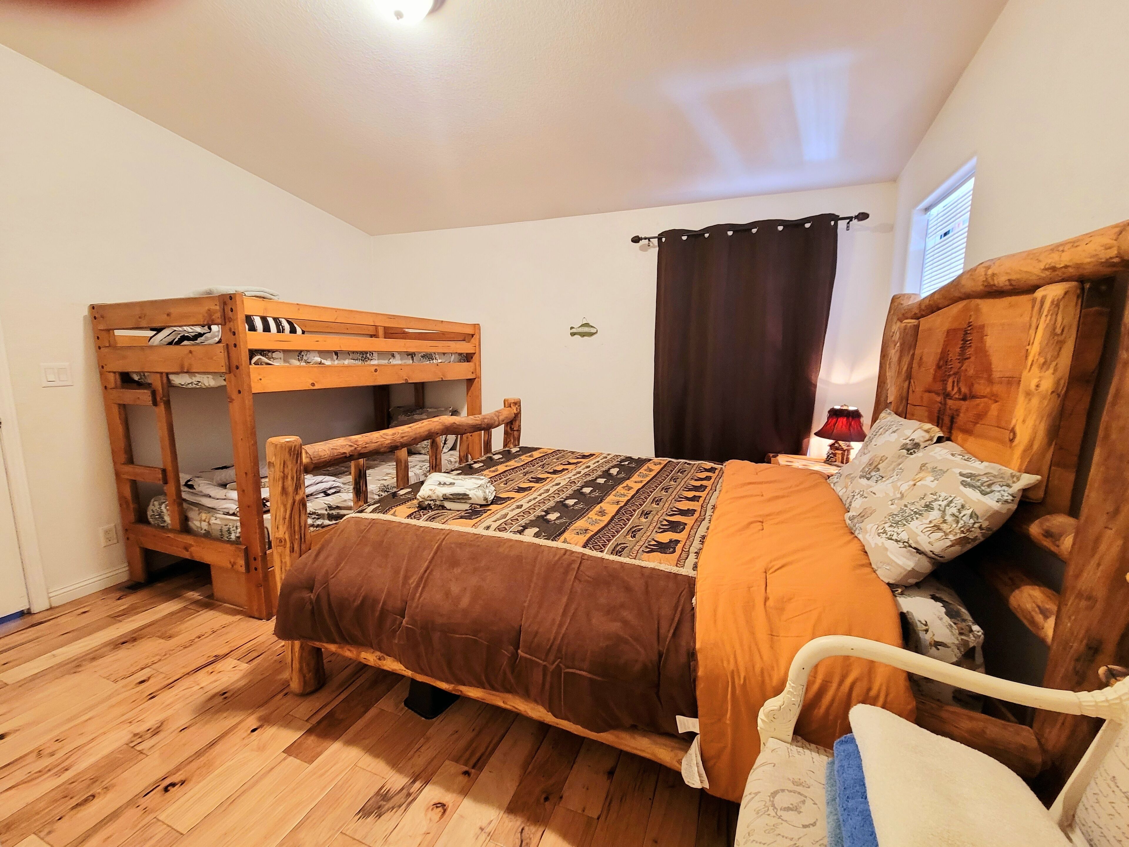 Una habitación con una litera, una cama individual con ropa de cama estampada, suelo de madera, una ventana con cortinas y una mesita con una lámpara.
