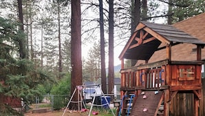 A wooden playhouse with a slide and climbing wall, surrounded by tall trees and a gravel path. - Myrtle Chalet (Big Bear City)