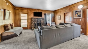 A cozy cabin interior with a stone fireplace, a large gray sofa, and wooden walls. - Myrtle Chalet (Big Bear City)