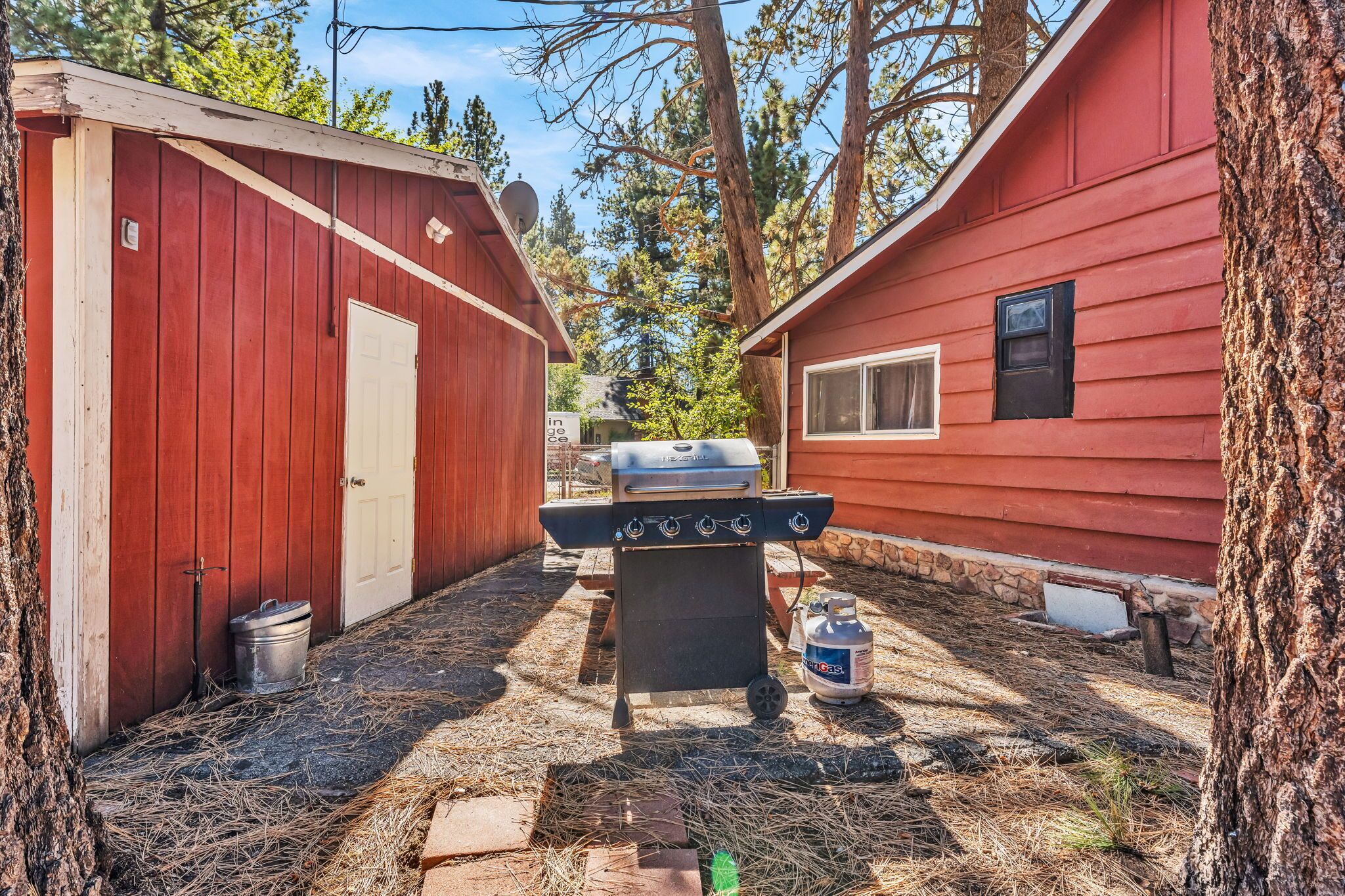 A red wooden building with a barbecue grill in the center, a gas canister nearby, and a stone foundation.