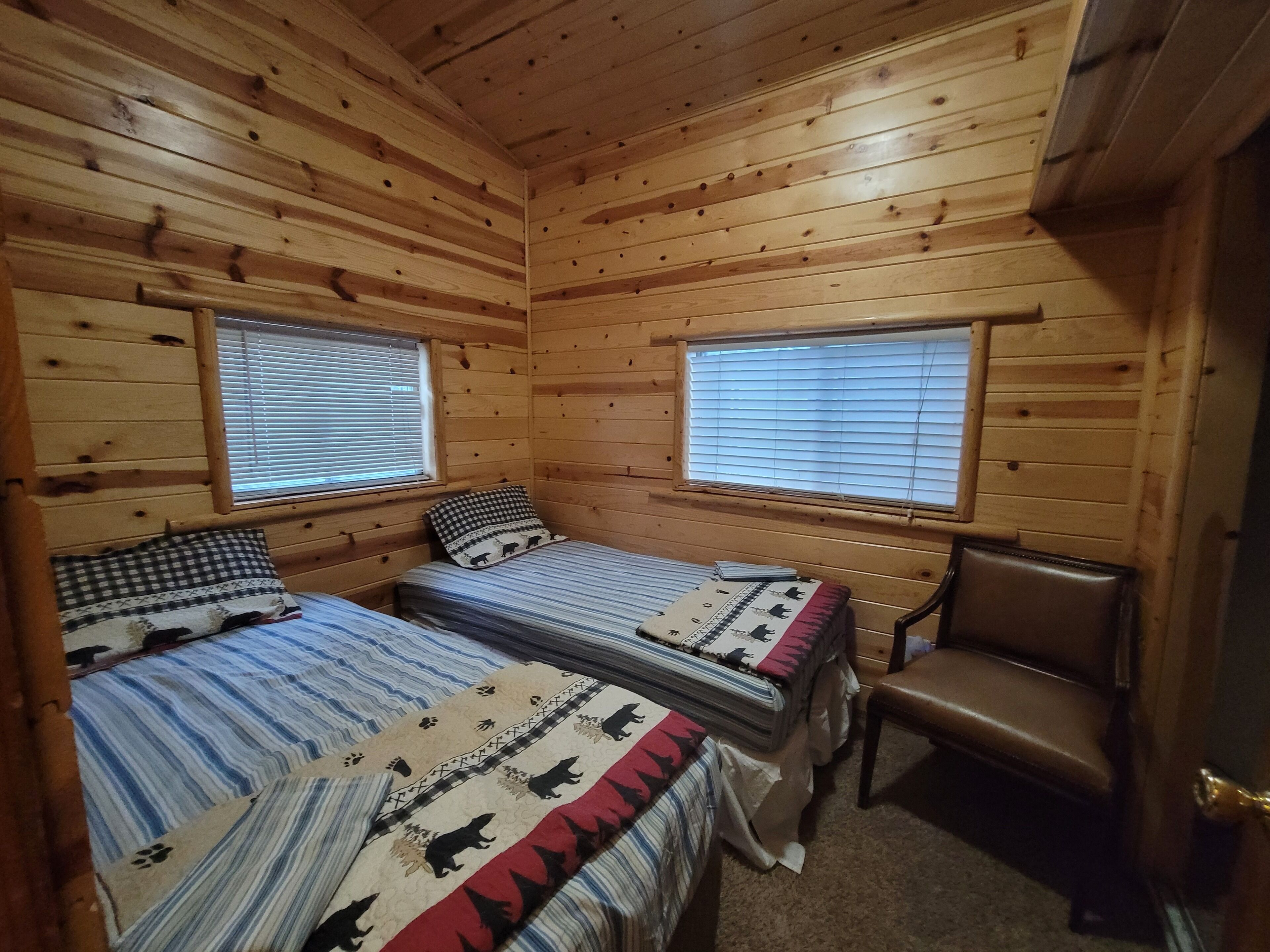 Une chambre de style cabane avec deux lits, une chaise et deux fenêtres équipées de stores.