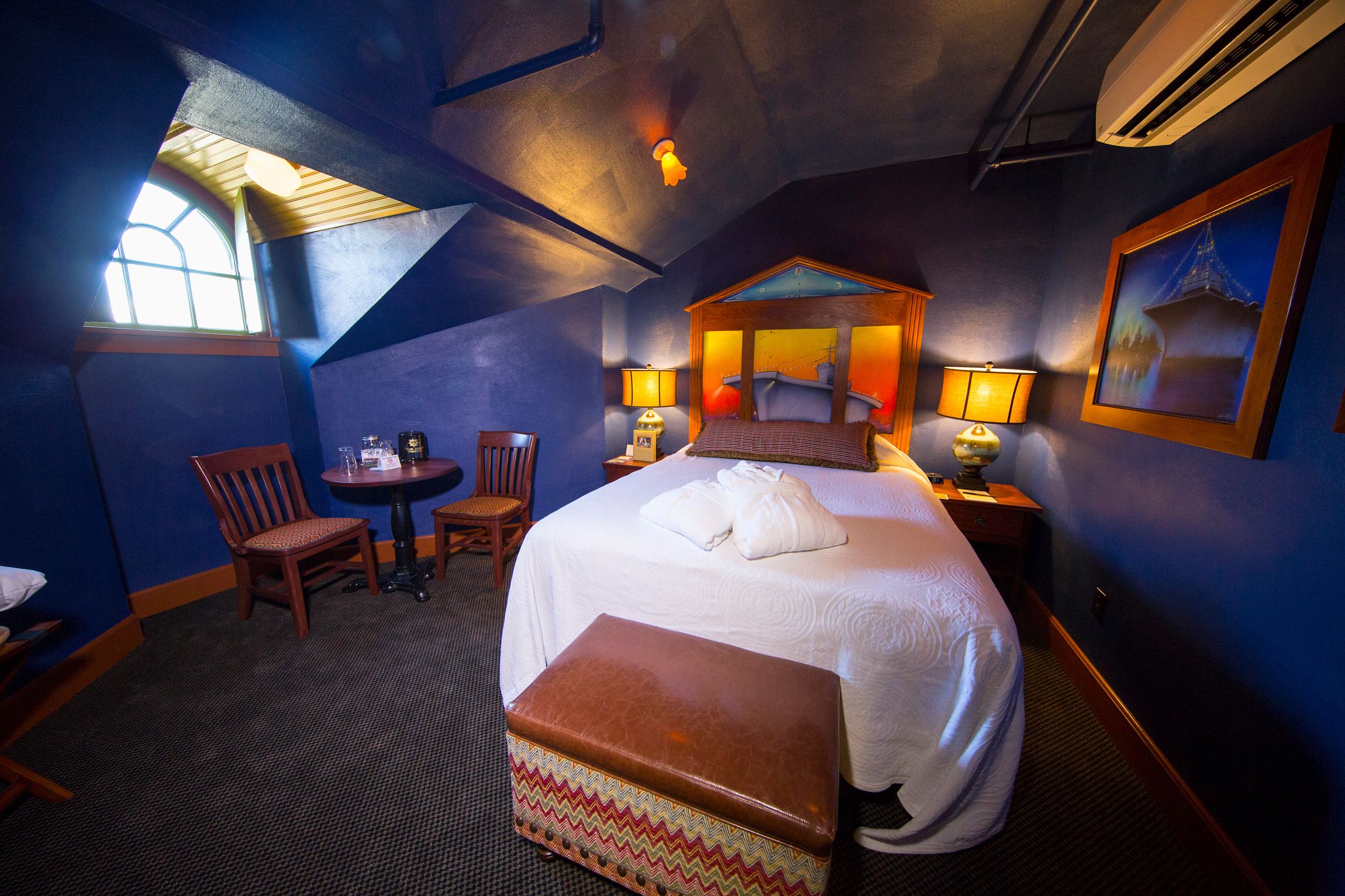 A hotel room with a large bed, a sitting area with a chair and a small table, and a window providing natural light.
