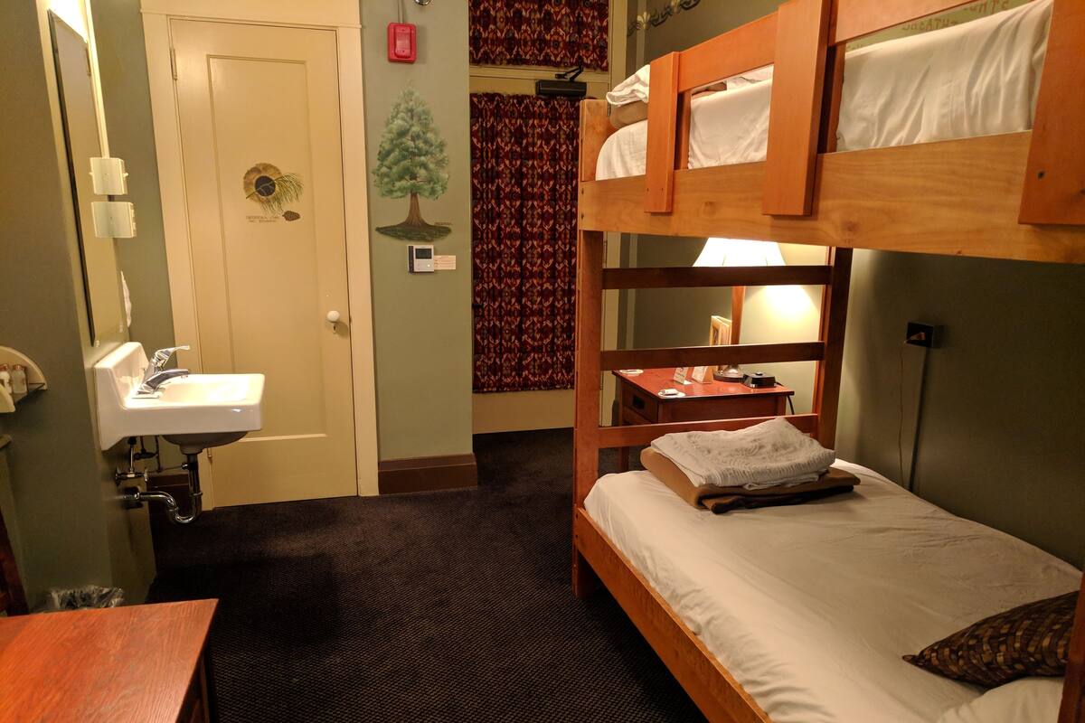 A compact hotel room with a bunk bed, a single bed, a small desk with a lamp, a wall-mounted telephone, and a bathroom with a sink and mirror.
