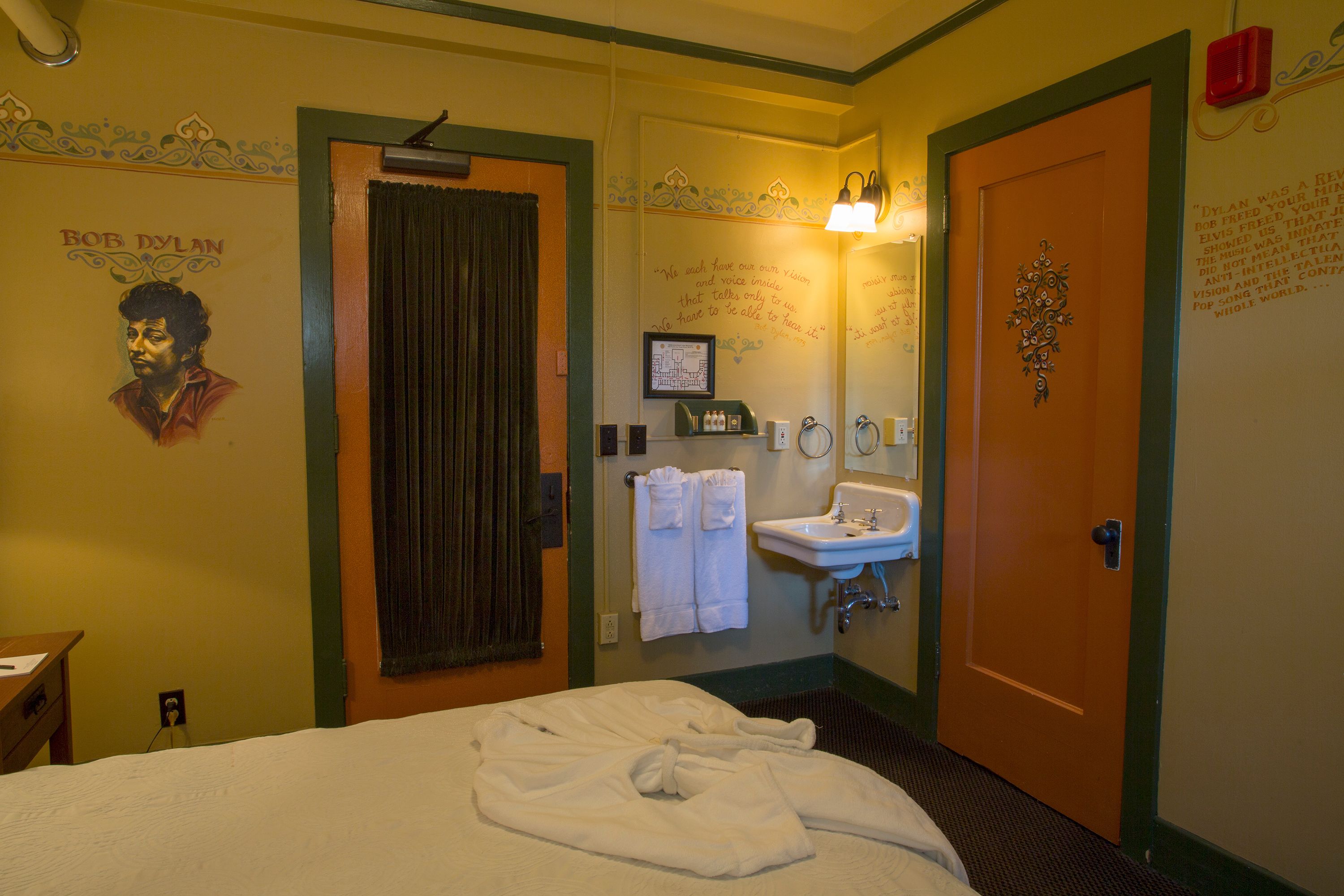 Een hotelkamer met een bed, een badkamer met een wastafel en spiegel, en een deur met decoratieve handgreep.