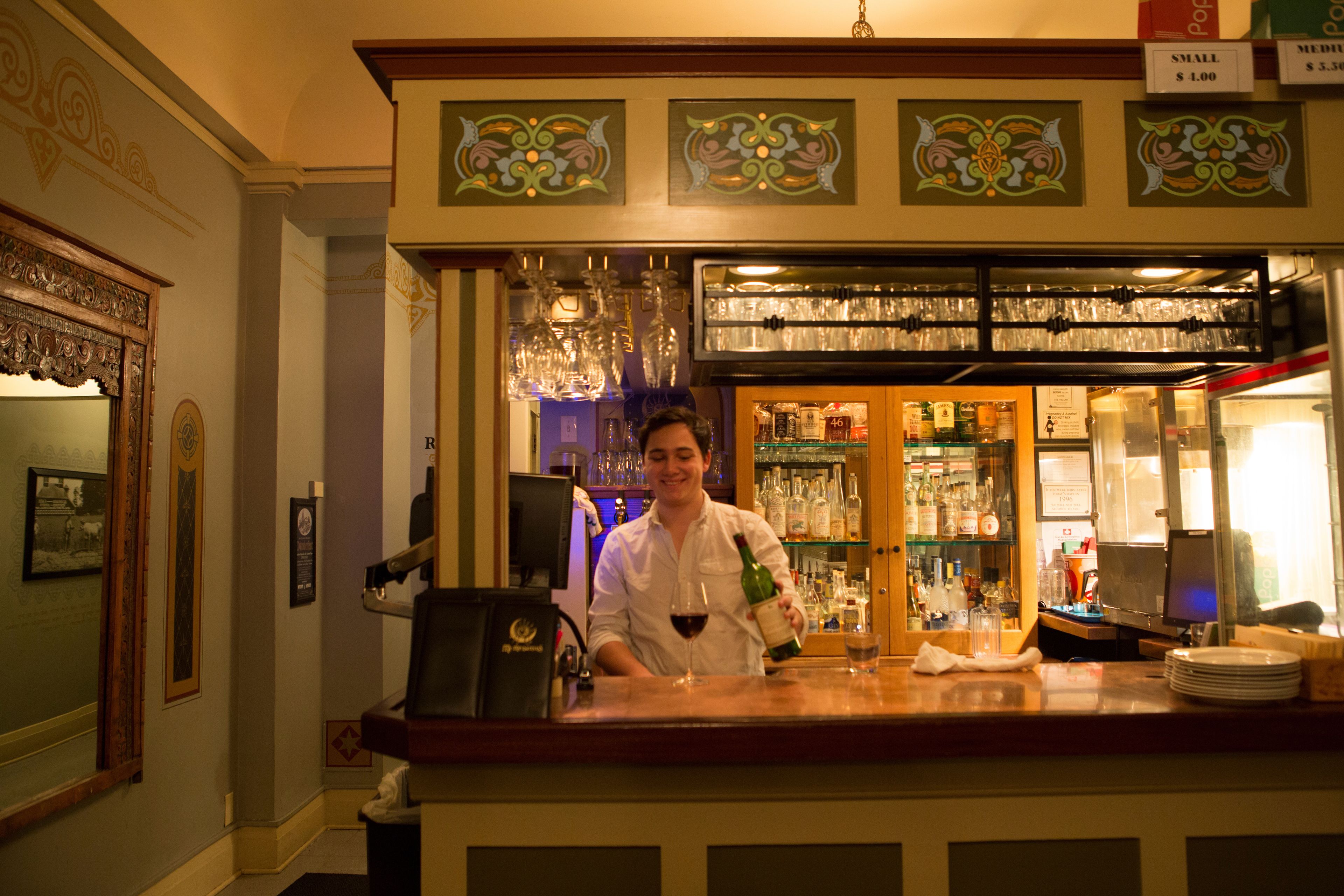 A bar area with a bartender, shelves with bottles, and a wine glass on the counter.