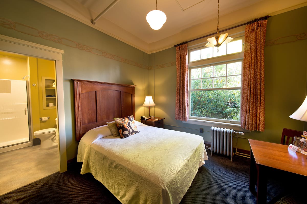 A hotel room with a bed, a wooden headboard, a nightstand with a lamp, a window with curtains, and a view of greenery outside.
