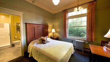Habitación de hotel con cama, mesita de noche, mesa, ventana con cortinas, radiador y puerta que da a un baño.