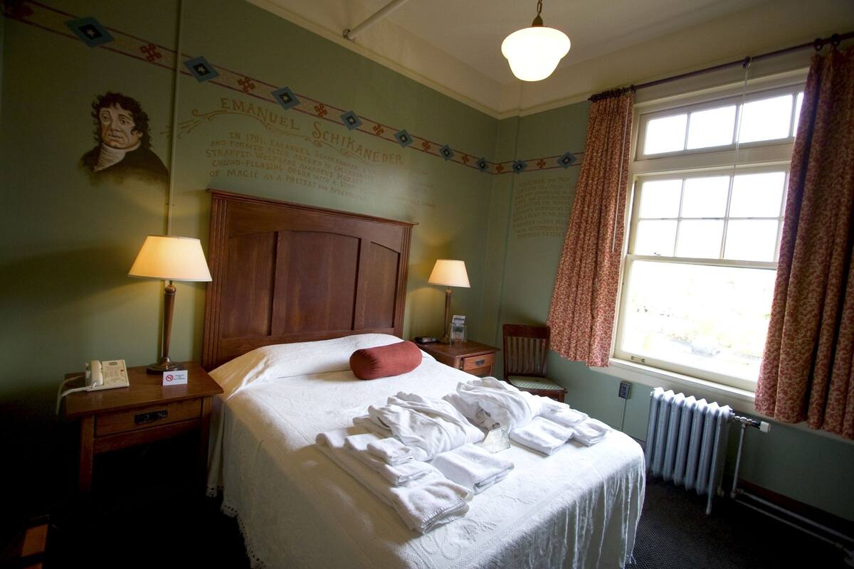 A hotel room with a large bed, two bedside tables with lamps, a window with curtains, and a wall with a portrait and text.