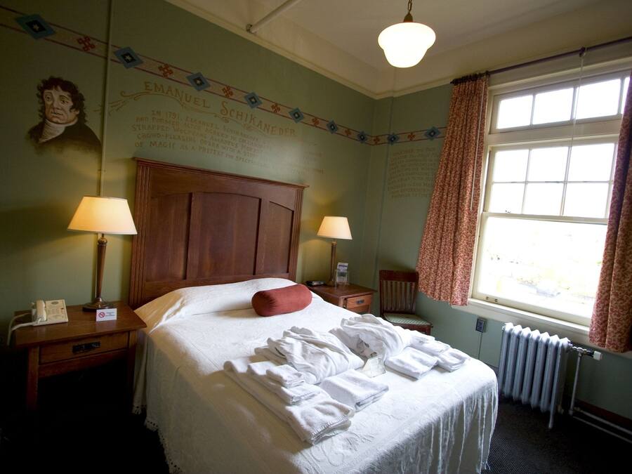 A hotel room with a large bed, two bedside tables with lamps, a window with curtains, and a wall with a portrait and text.
