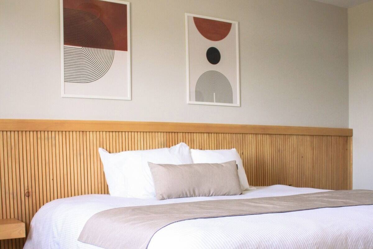 A bedroom with a wooden headboard, a bed with white bedding and a beige throw, and two abstract wall art pieces.