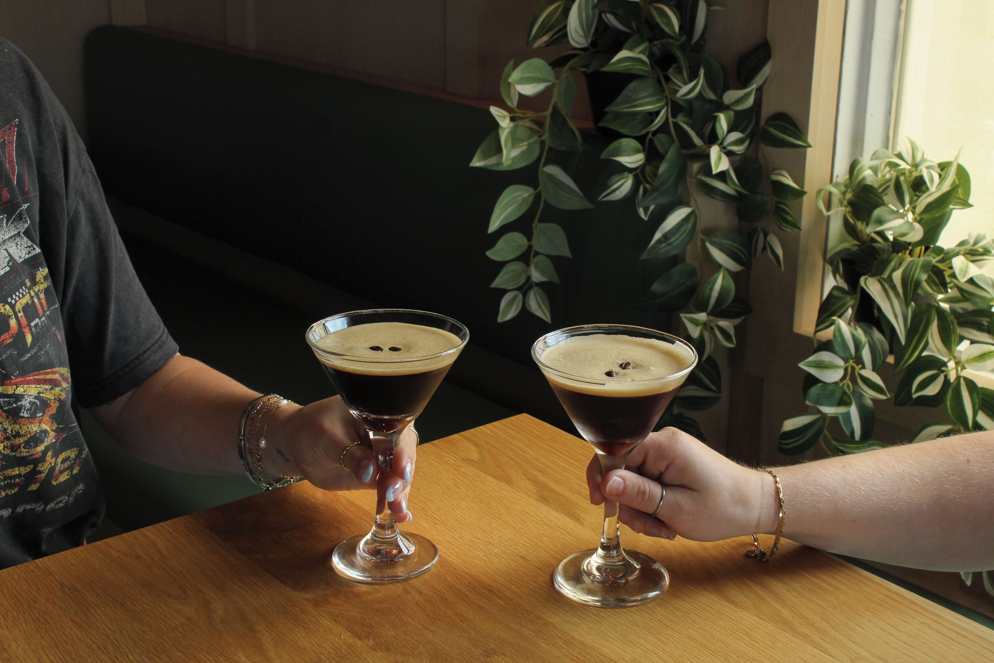Two people seated at a wooden table with drinks in martini glasses, a plant in the background, and a dark-colored couch.