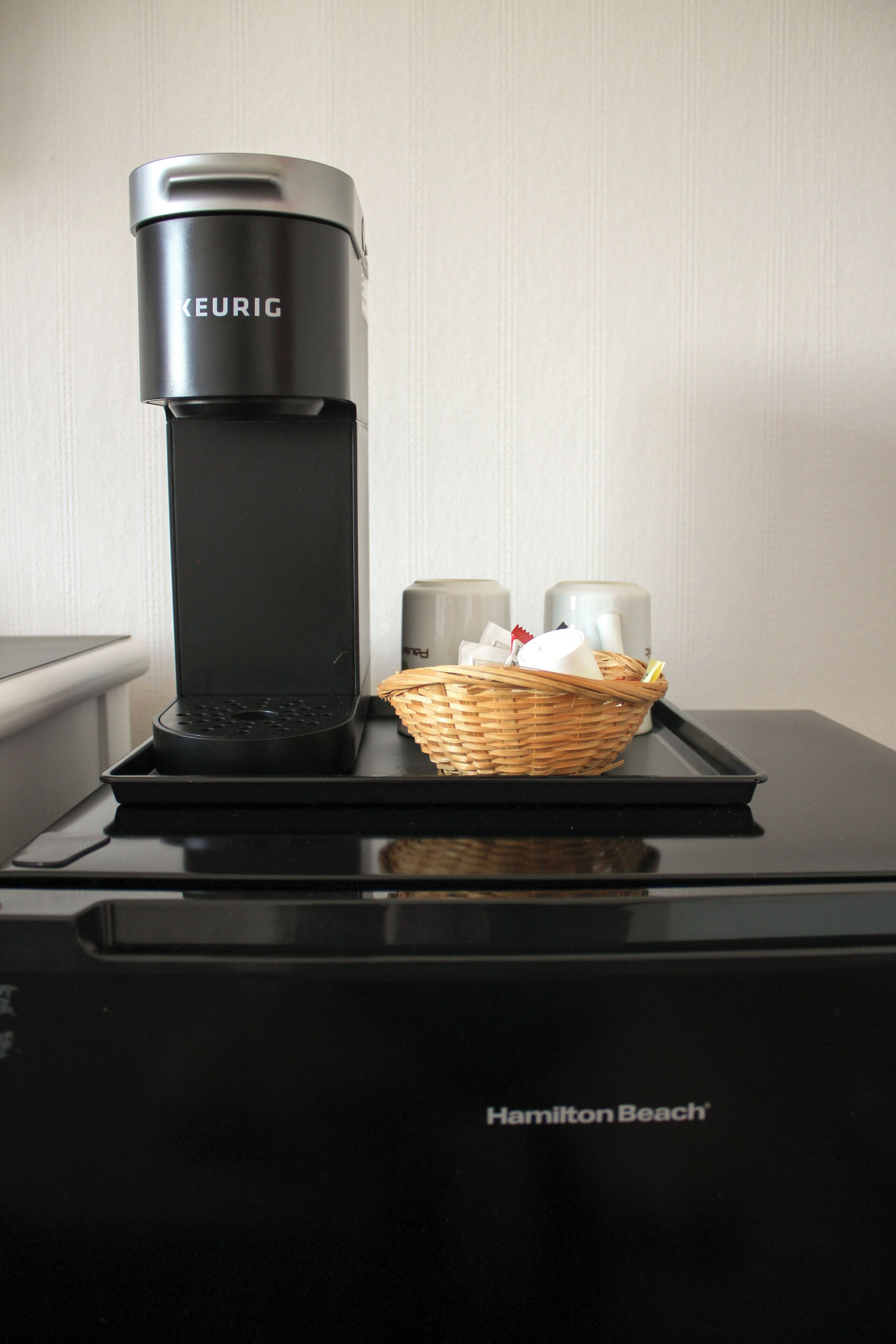 A Keurig coffee maker on a Hamilton Beach appliance with a basket of items on the tray.