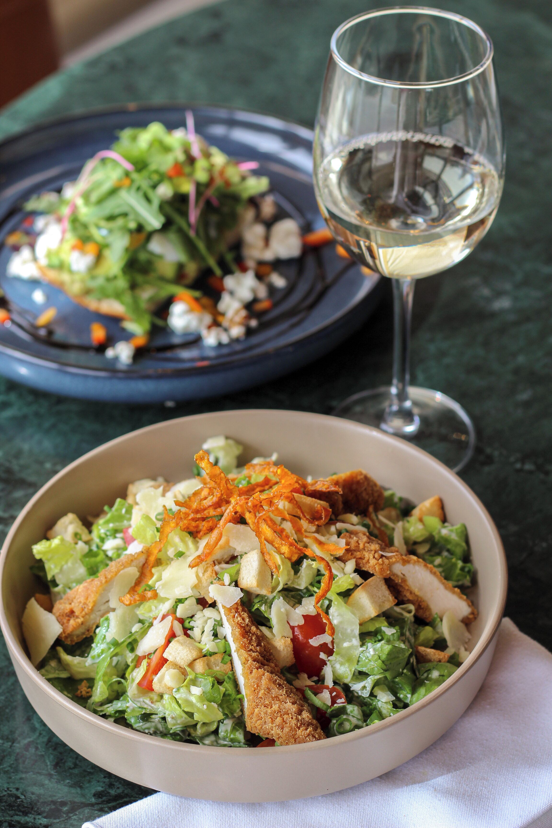Een saladekom met croutons en geraspte kaas, vergezeld van een glas witte wijn op een tafel.