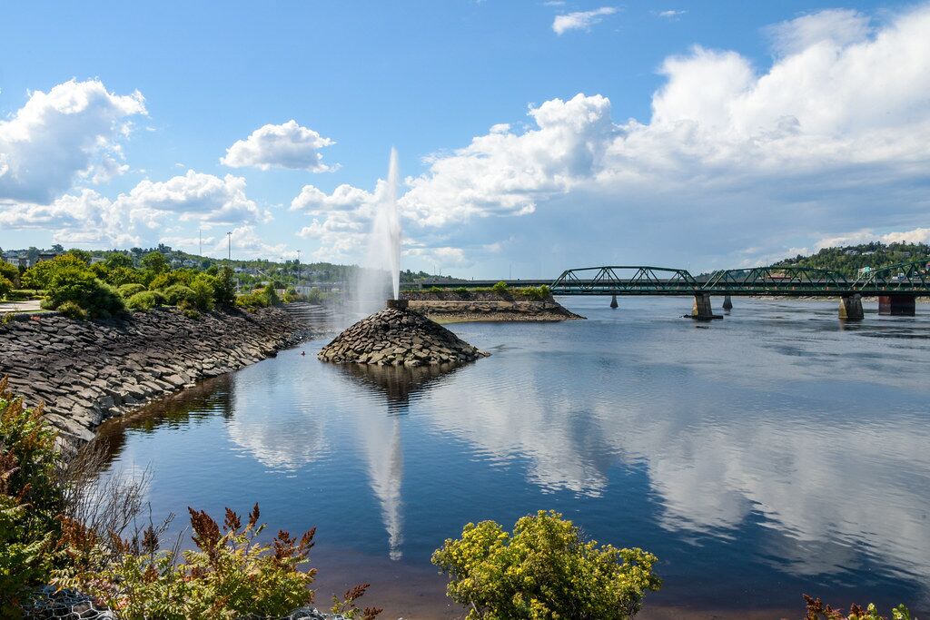 A calm river with a fountain, a stone jet, and a bridge in the background.