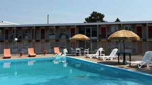 Una piscina con sillas de descanso y sombrillas, un edificio con varios balcones y un cielo despejado.