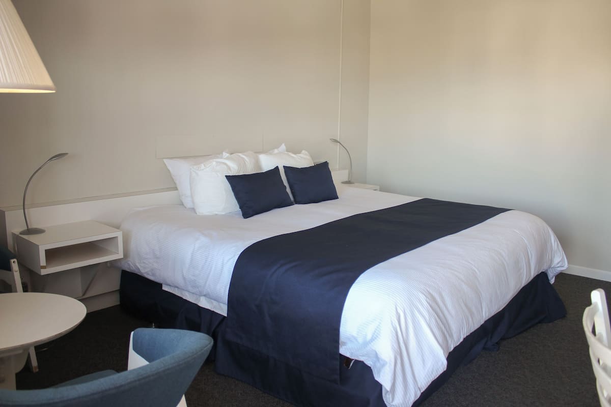 A hotel room with a neatly made bed, a small round table, a chair, and a bedside lamp.