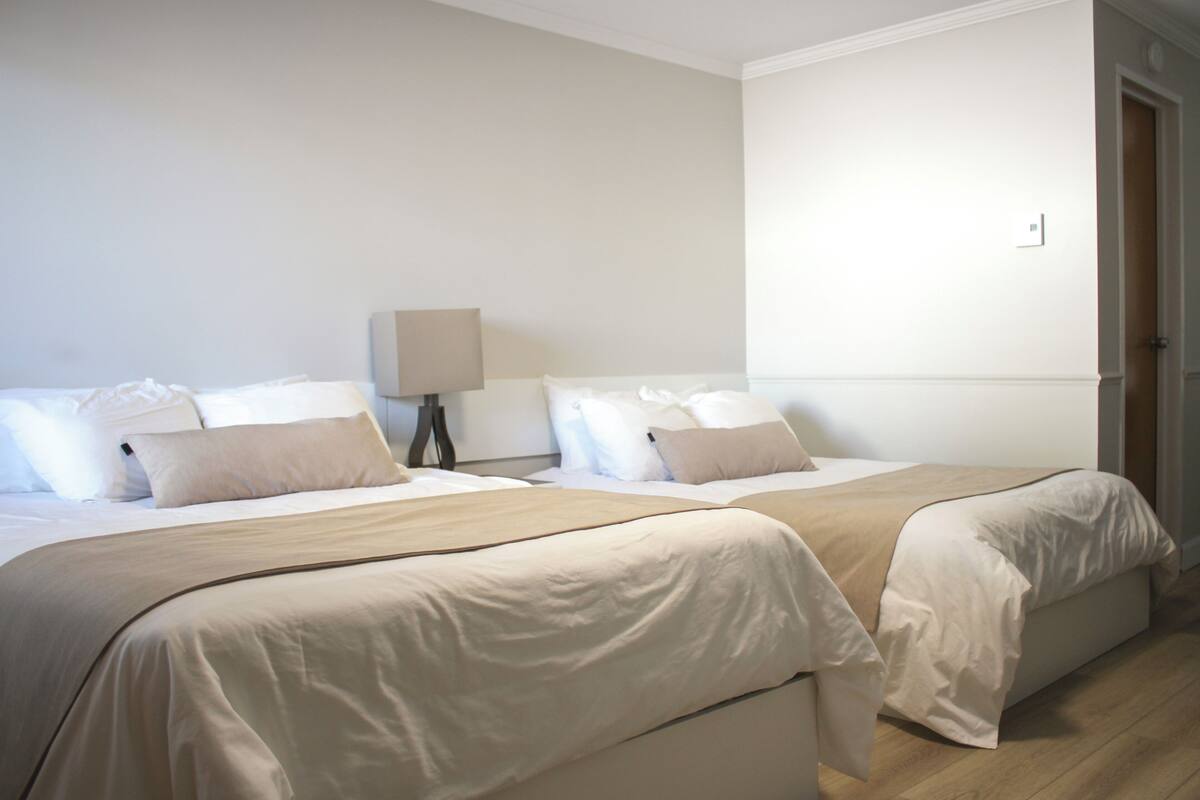 A hotel room with two beds, a nightstand with a lamp, and a door.