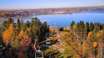 Een skilift met gondels die een helling oprijden, omgeven door herfstkleuren en een uitzicht op een meer en een stad in de verte.