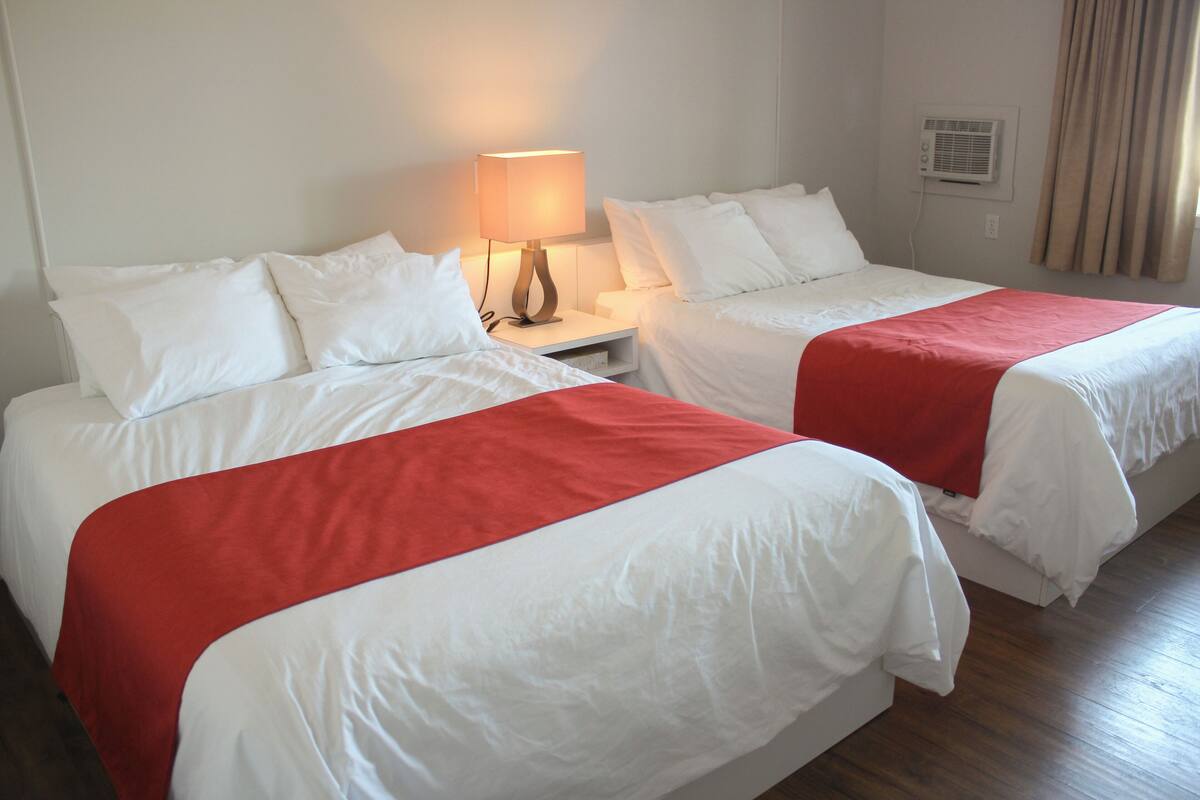 A hotel room with two beds, each with white bedding and red bedspreads. There is a bedside table with a lamp between the beds.