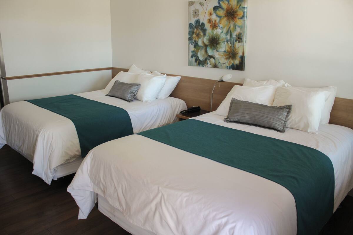 A hotel room with two beds, each with white bedding and a teal throw blanket. There is a floral artwork on the wall above the beds.