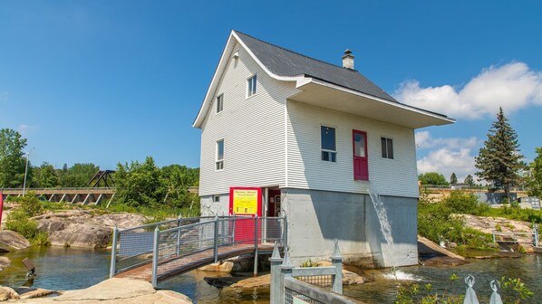 A small white building with a red door, situated by a water body with a bridge leading to it. - Auberge Le Parasol  (Saguenay)