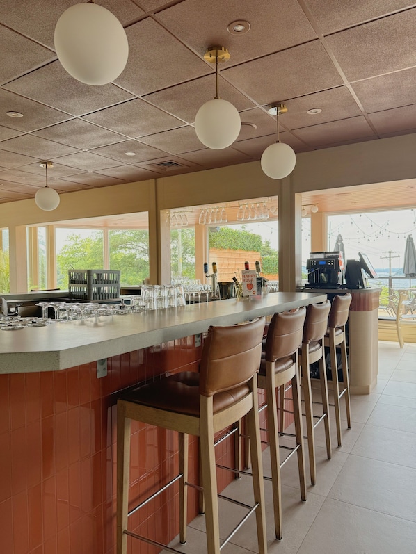 A bar area with a long counter, multiple stools, hanging light fixtures, and a view of the outdoors. - Auberge Le Parasol  (Saguenay)
