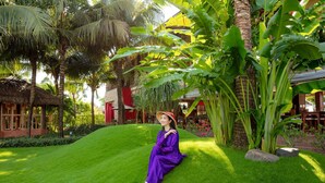 A person sitting on a well-manicured lawn with tropical plants and a thatched-roof structure in the background. - The Island Lodge (Thoi Son)
