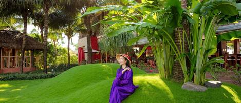 A person sitting on a well-manicured lawn with tropical plants and a thatched-roof structure in the background.