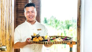 A person holding a tray with various food items, standing in front of a wooden door with a view of greenery outside. - The Island Lodge (Thoi Son)