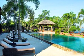 A pool area with lounge chairs, a thatched-roof gazebo, and palm trees.