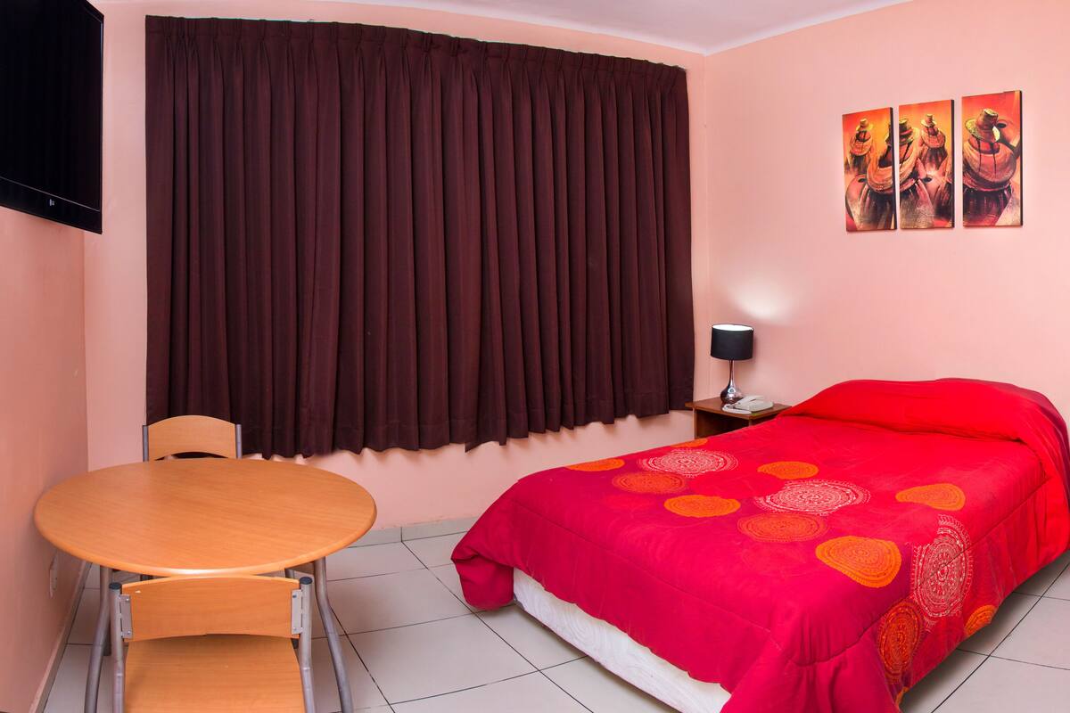 A hotel room with a bed, a table, a chair, a TV, and artwork on the wall.