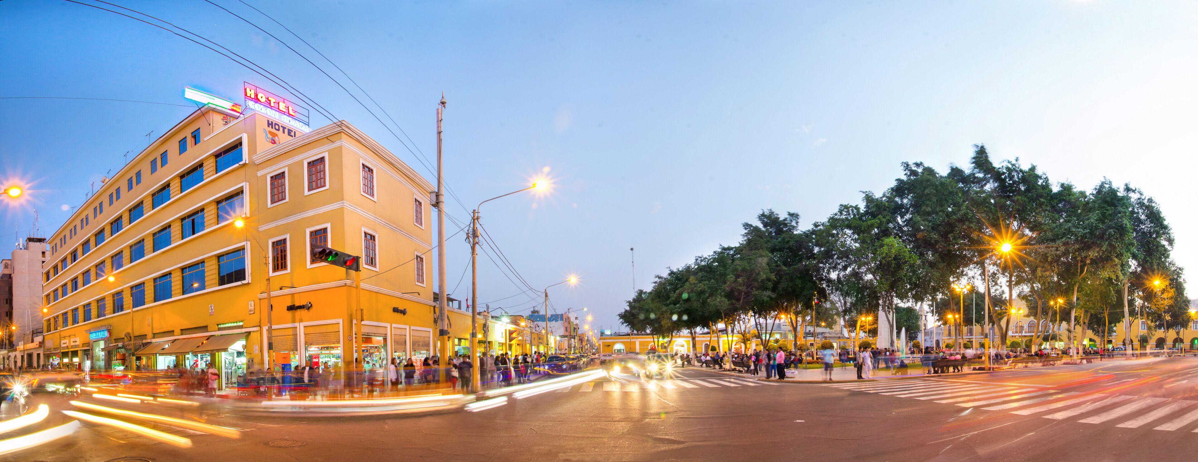 Un hotel de varios pisos con un llamativo letrero 'HOTEL', un paso de peatones y una calle bien iluminada con árboles y bancos.