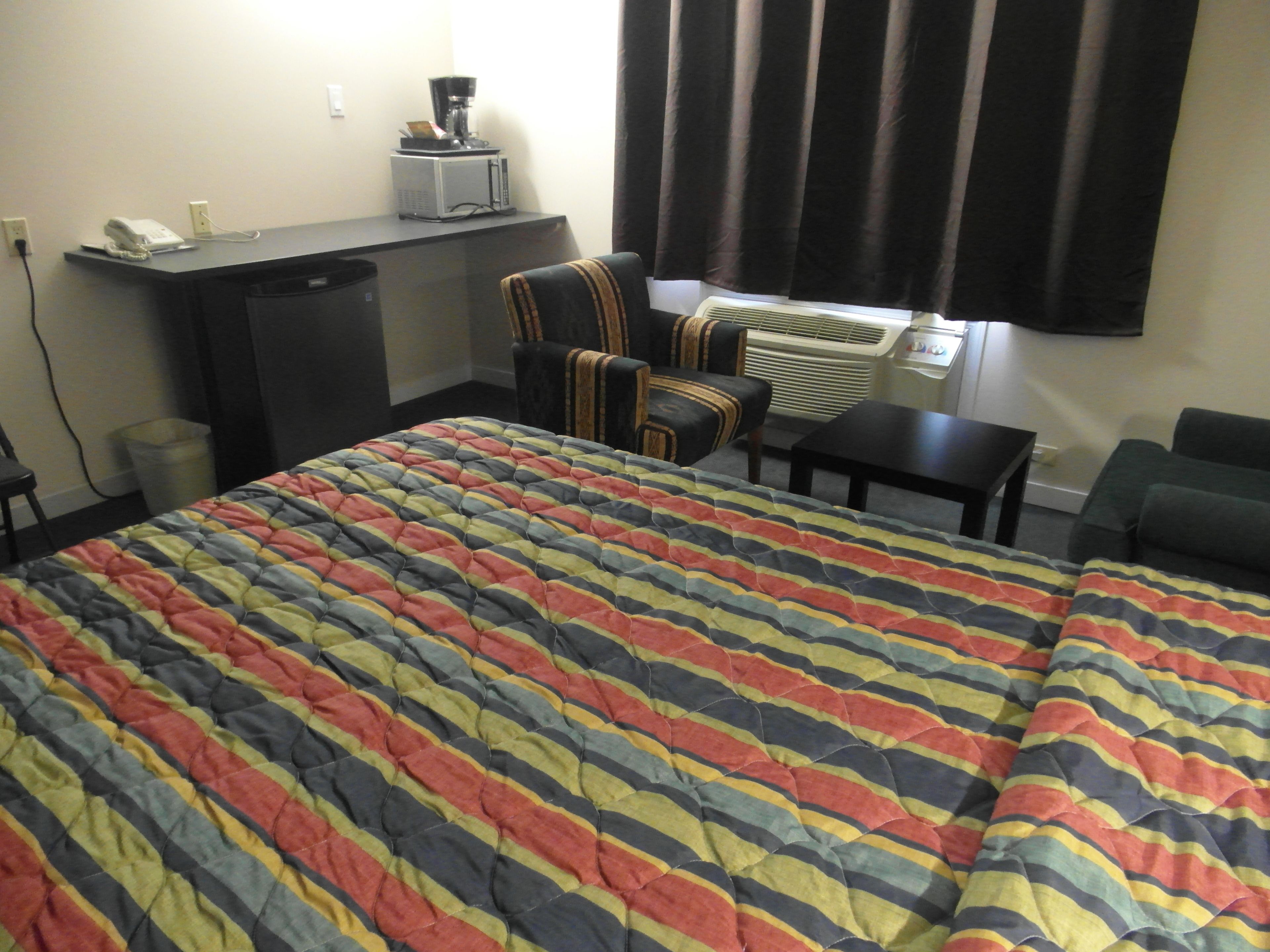 A hotel room with a large bed, a desk with a coffee maker, a chair, an air conditioning unit, and a window with curtains.