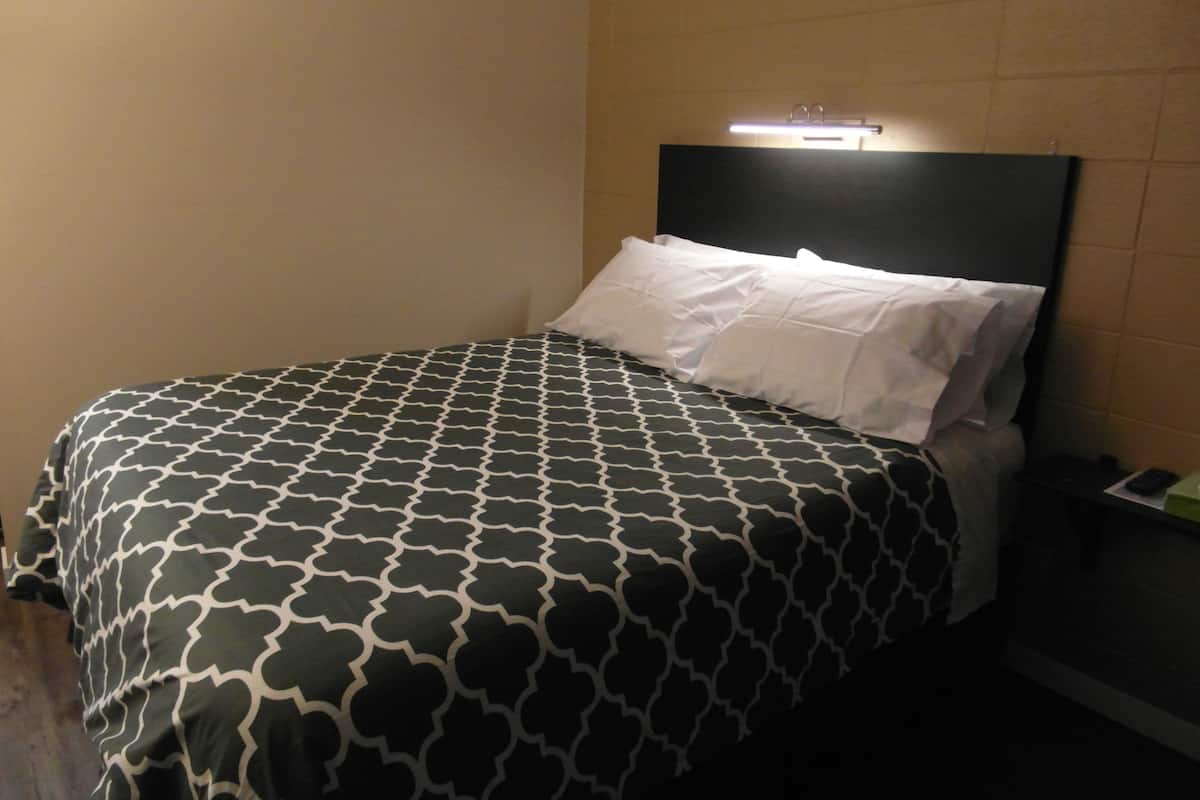 Ett hotellrum med en säng, en sänggavel med en lampa och ett nattduksbord med en telefon och ett grönt föremål.