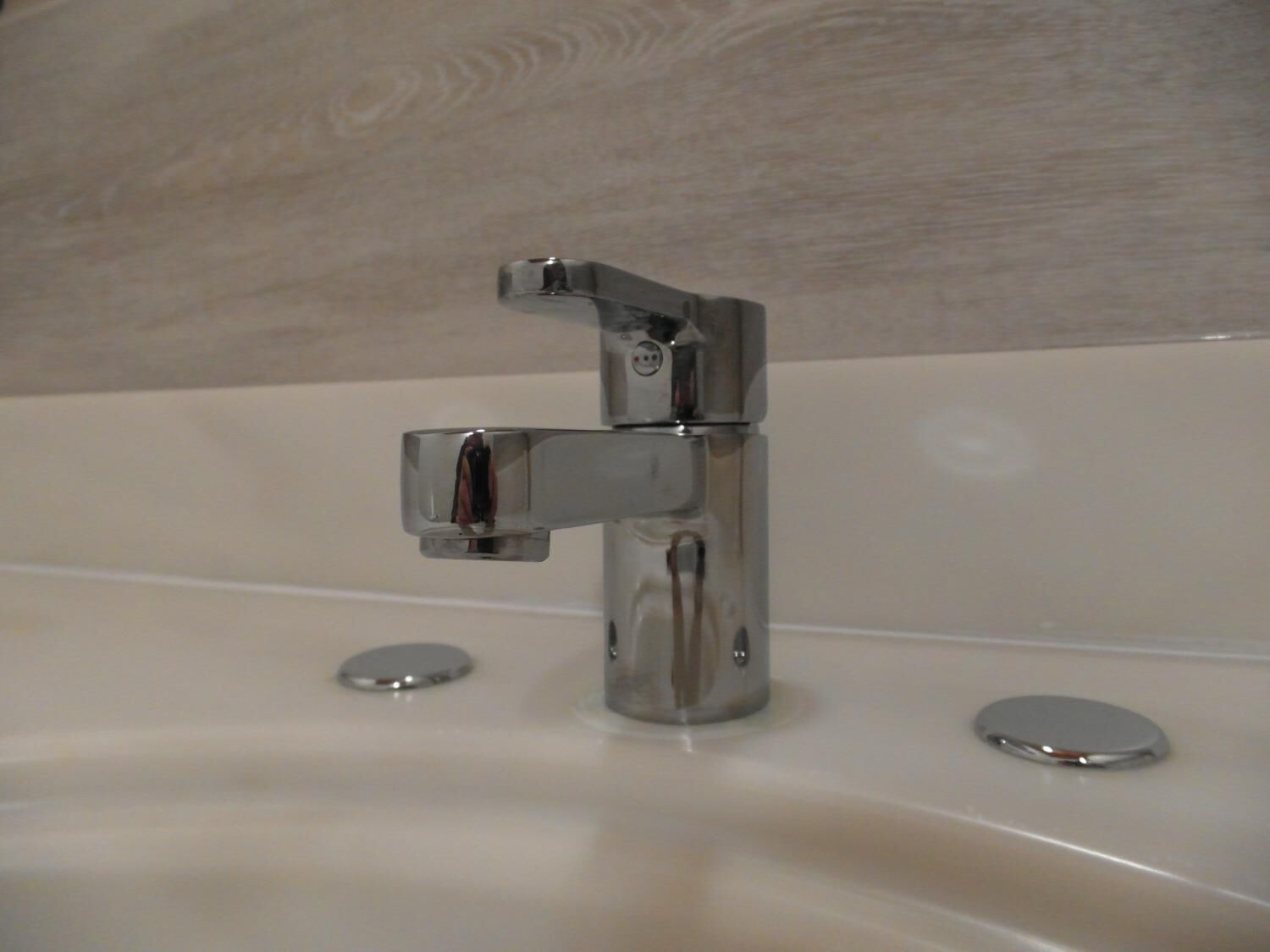 A chrome faucet mounted on a white bathroom sink with a wooden backsplash.