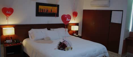 Habitación de hotel con una cama grande, dos mesitas de noche con lámparas, un cuadro enmarcado en la pared y un ramo de flores sobre la cama.