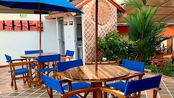 Un patio al aire libre con mesas de madera y sillas azules, cubierto por sombrillas azules y rodeado de plantas y un muro decorativo.