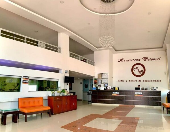 A hotel lobby with a reception desk, a chandelier, a seating area with a sofa and chairs, and a large window with a view. - Hotel Hacaritama Colonial (Villavicencio)