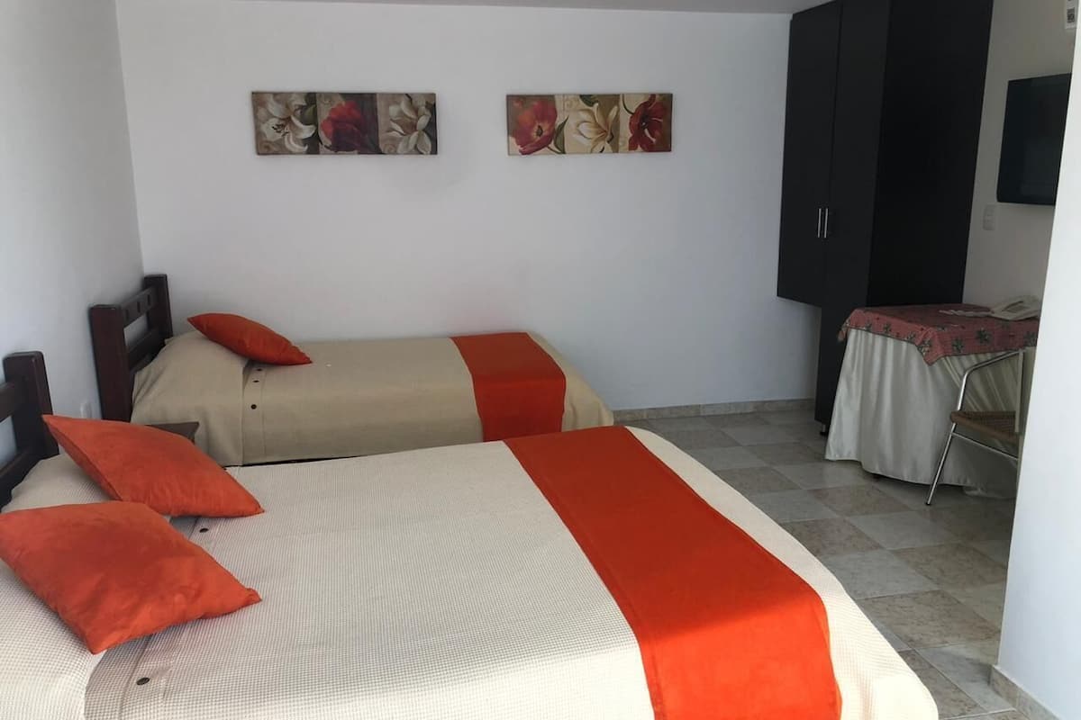 Ein Zimmer mit zwei Betten, jeweils mit einer roten und beigen Bettdecke, und einem dunklen Kleiderschrank. Darüber hängen zwei an der Wand befestigte Bilder.