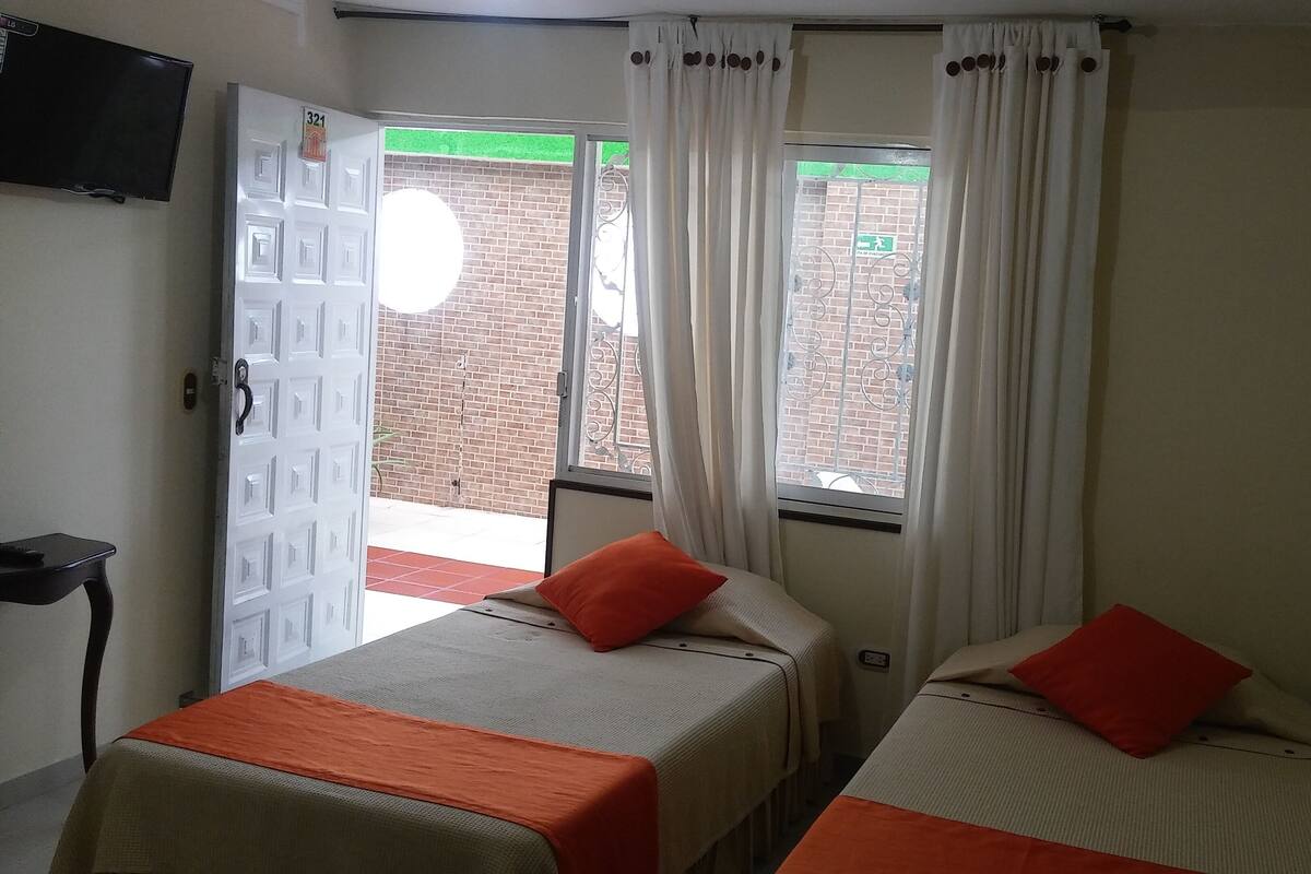Ein Zimmer mit zwei Betten, einem Fernseher, einer Tür und einem fensterbepflanzten Fenster.