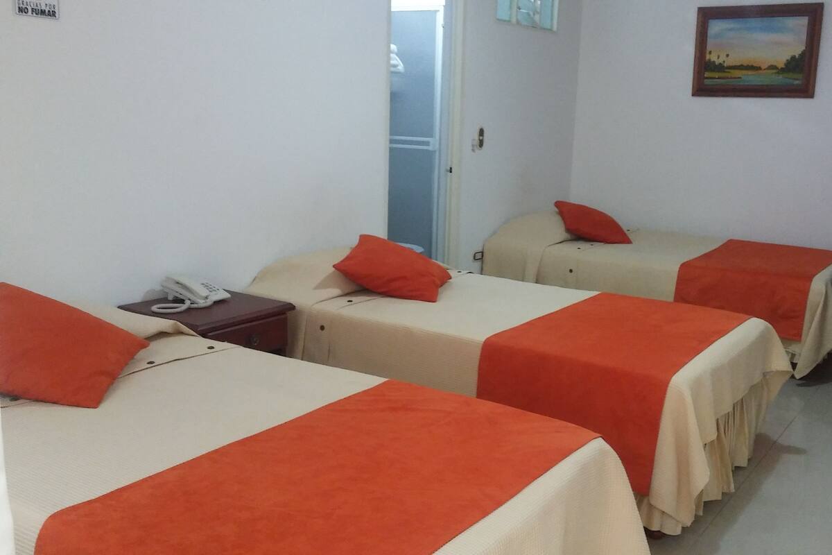 Ein Hotelzimmer mit drei Betten, jedes mit einer roten Decke und einer beigefarbenen Tagesdecke. Ein Nachttisch mit Telefon und Lampe ist vorhanden. Ein Gemälde hängt an der Wand, und im Hintergrund ist eine Tür zu sehen.