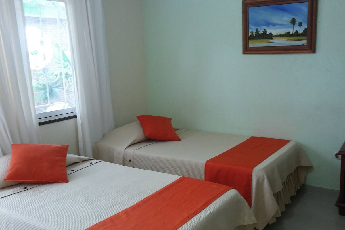 Ein Zimmer mit zwei Betten, jedes mit einem roten Kopfkissen und einer weißen Tagesdecke. An der Wand hängt ein Gemälde und es gibt ein Fenster mit Vorhängen.