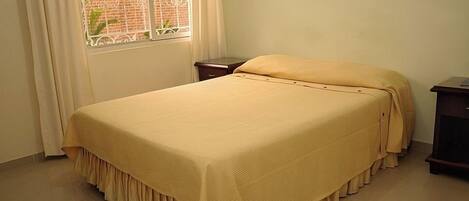 A single bed with a yellow bedspread in a room with a window and a small bedside table.